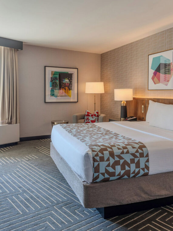 Slaapkamer in het Valley Forge Casino Resort met een kingsize bed en een geometrisch plaid, abstracte kunst boven het hoofdeinde en een balkon met uitzicht op beboste heuvels.