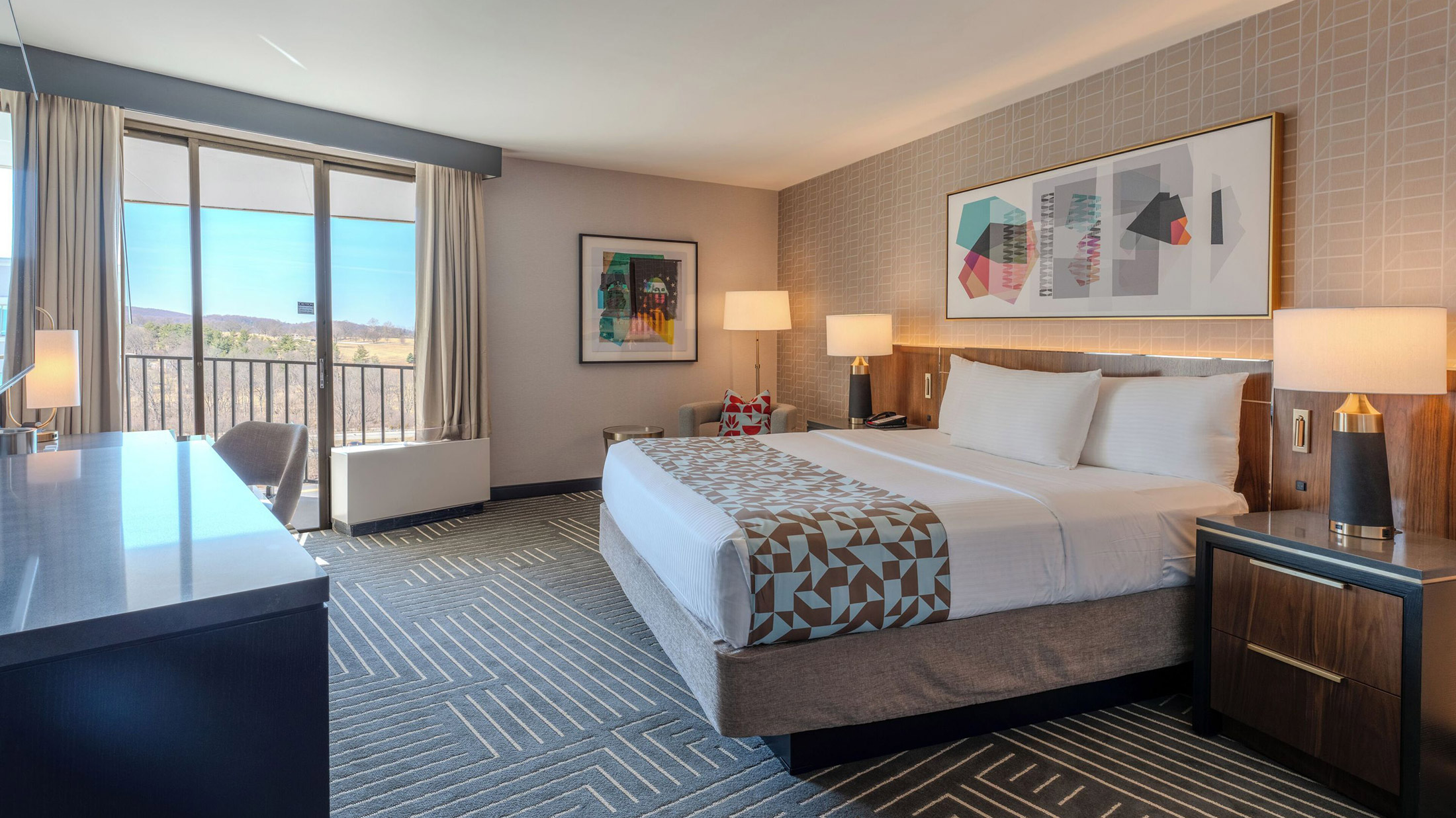 Chambre au Valley Forge Casino Resort, dotée d'un lit king size et d'un plaid à motifs géométriques, d'une œuvre d'art abstraite au-dessus de la tête de lit et d'un balcon donnant sur des collines boisées.