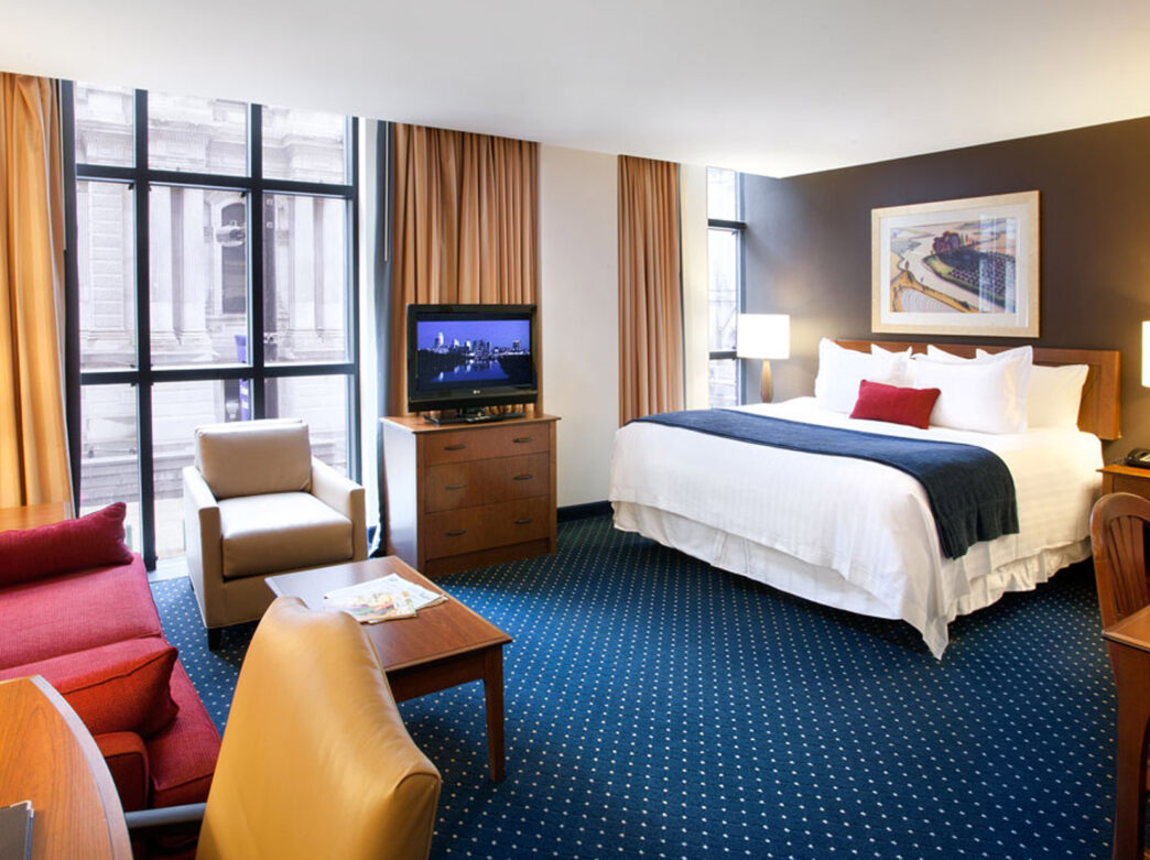 A spacious hotel room at the Residence Inn with a king-size bed, a red sofa, and a comfortable armchair. Large windows offer plenty of natural light and a television sits on the dresser. The room is decorated in warm colors with a blue carpet.