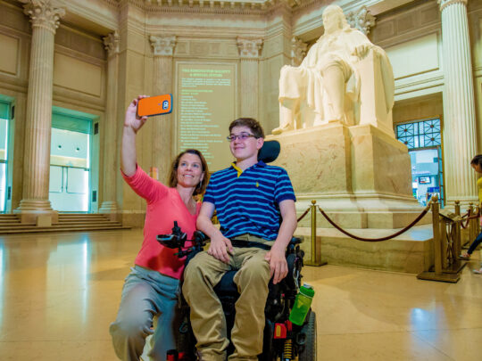 Un joven visitante en silla de ruedas posa para un selfie en el vestíbulo accesible The Franklin Institute