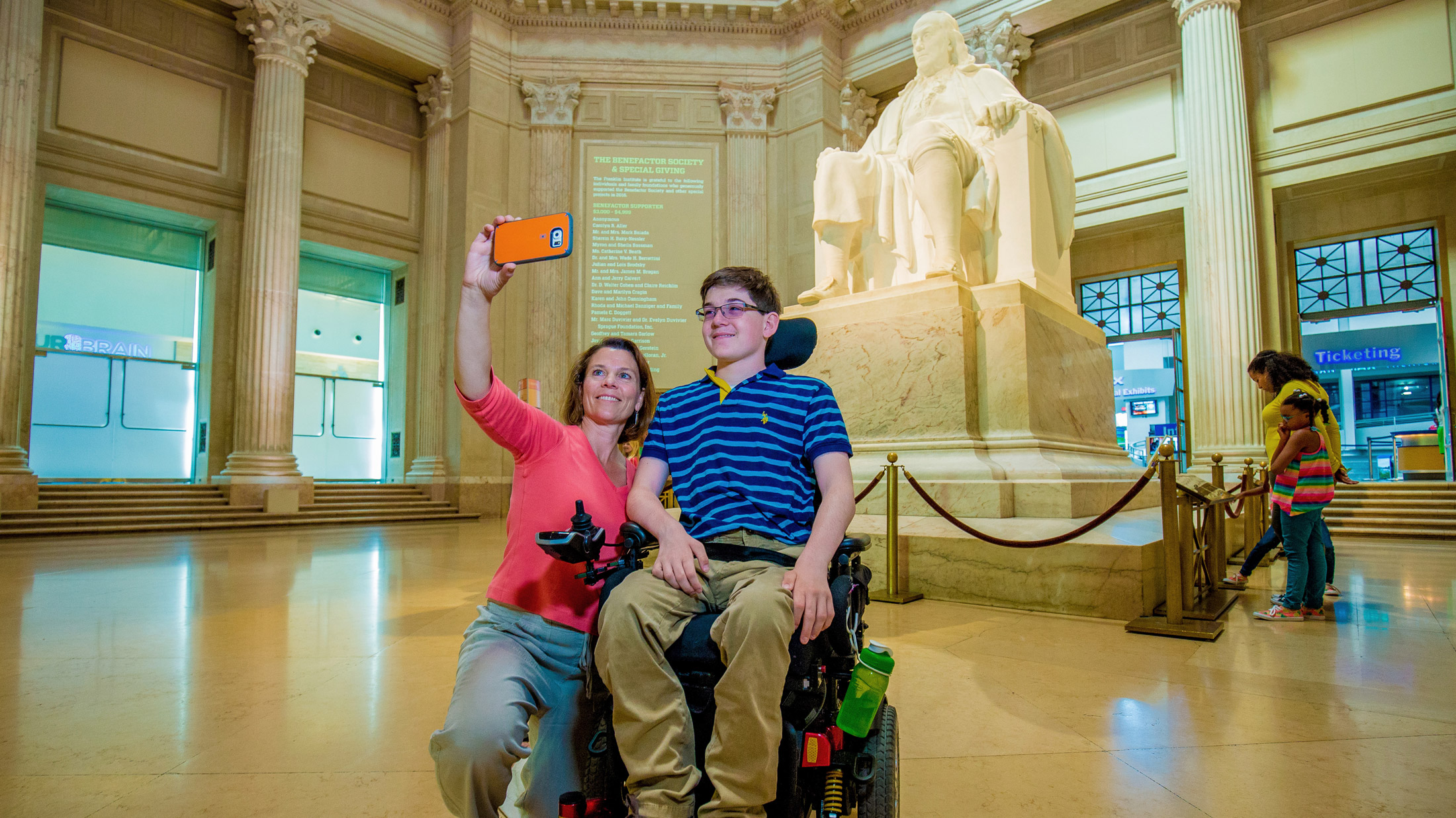 A young visitor using a wheelchair poses for a selfie in accessible The Franklin Institute lobby