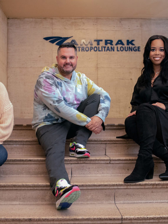 The hosts of the Love + Grit podcast sit on the steps in front of a sign for Amtrak