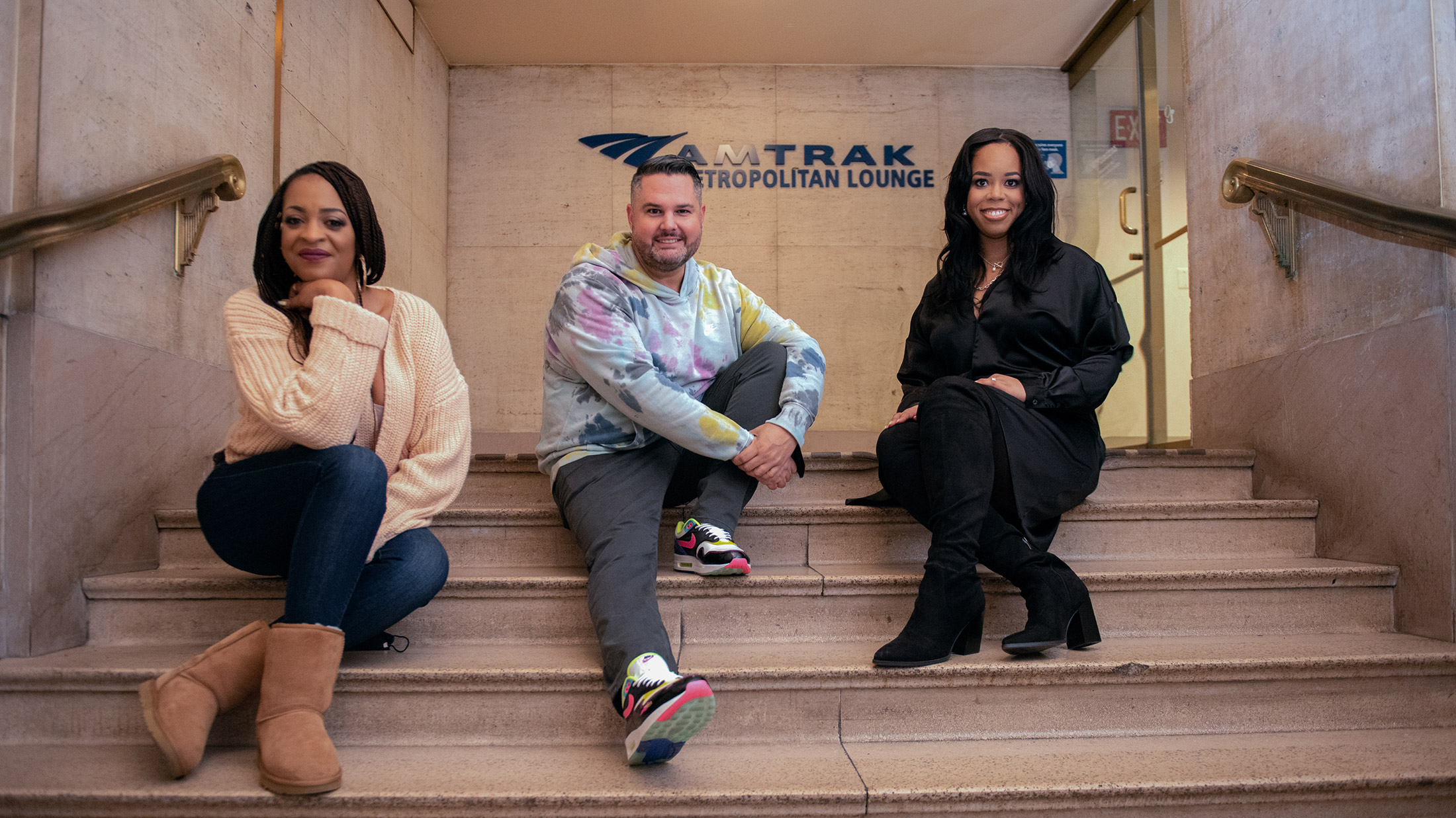 De presentatoren van de podcast Love + Grit zitten op de trap voor een bord van Amtrak.