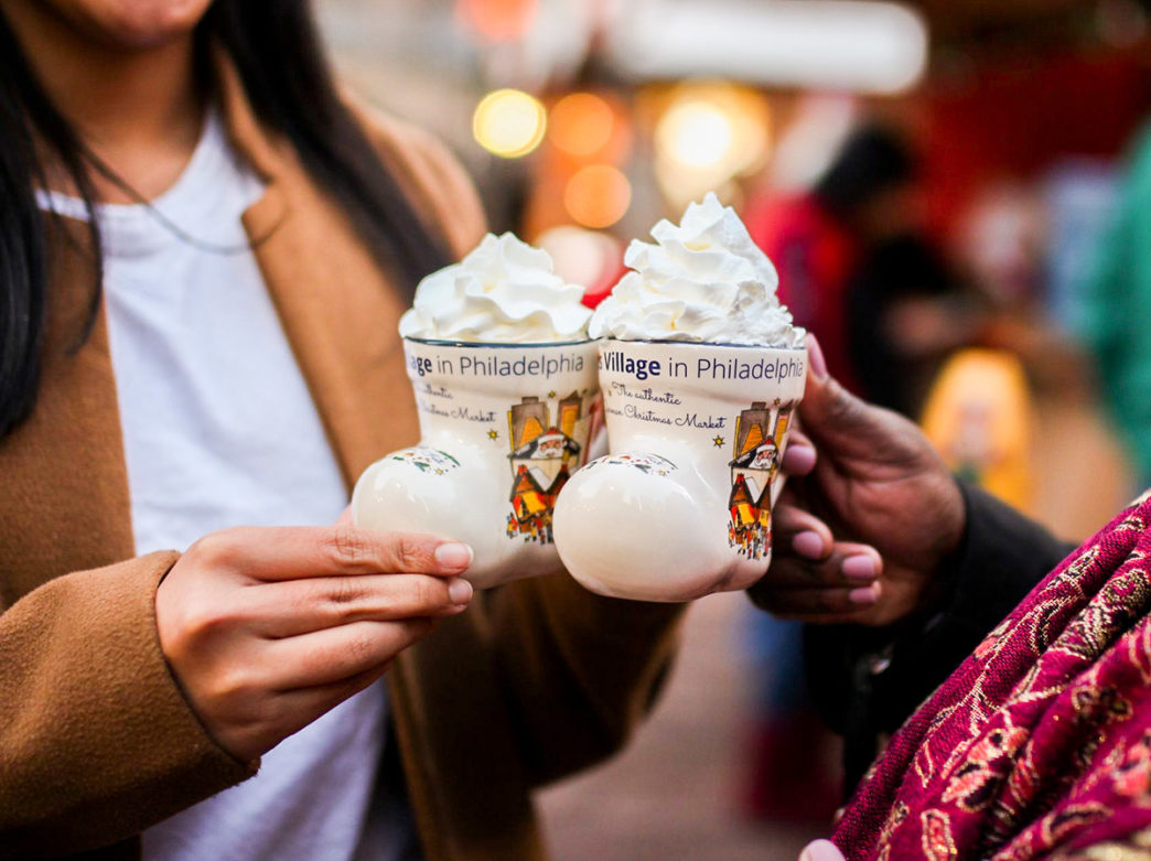 Two people toast mugs with whipped cream on the top at Christmas Village in Philadelphia