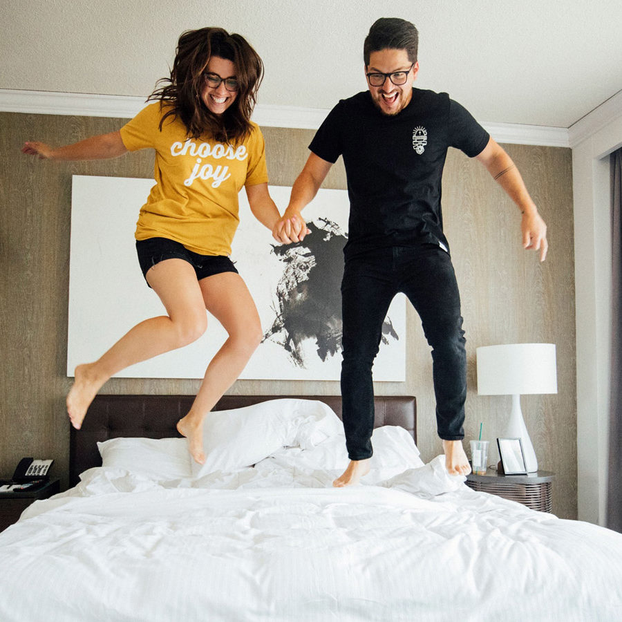 A woman in shorts and a T-shirt and a man in a T-shirt and jeans jump on the bed at The Logan, Philadelphia's Hotel
