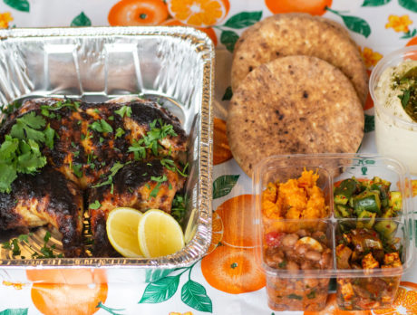 Takeout containers hold chicken topped with cilantro, an array of salads and toasted pita, resting on a table cloth covered in oranges