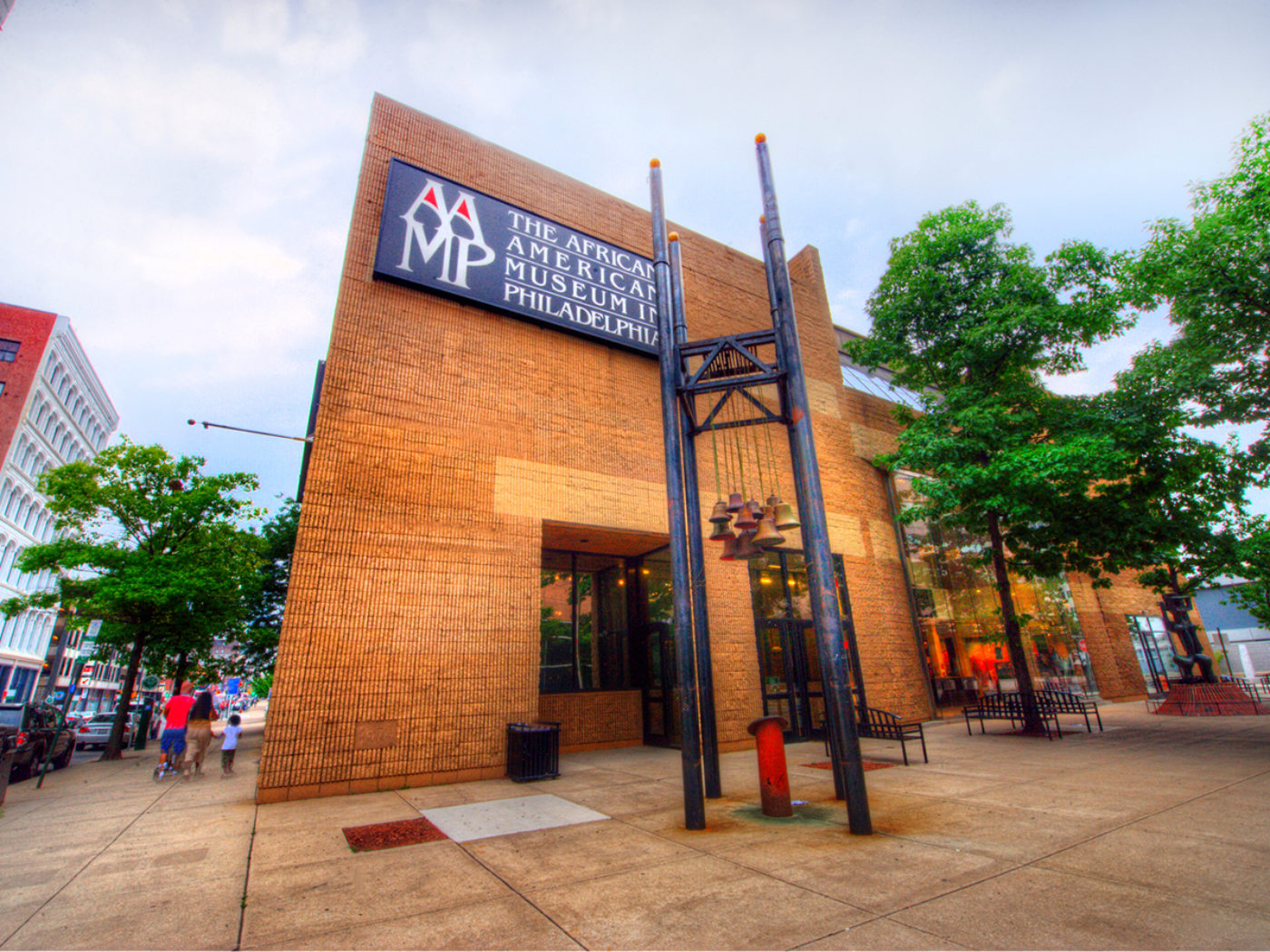 Exterior view of the African American Museum in Philadelphia, a brick building with a large sign on the facade and a sculptural bell structure in front, set along a city sidewalk lined with trees.