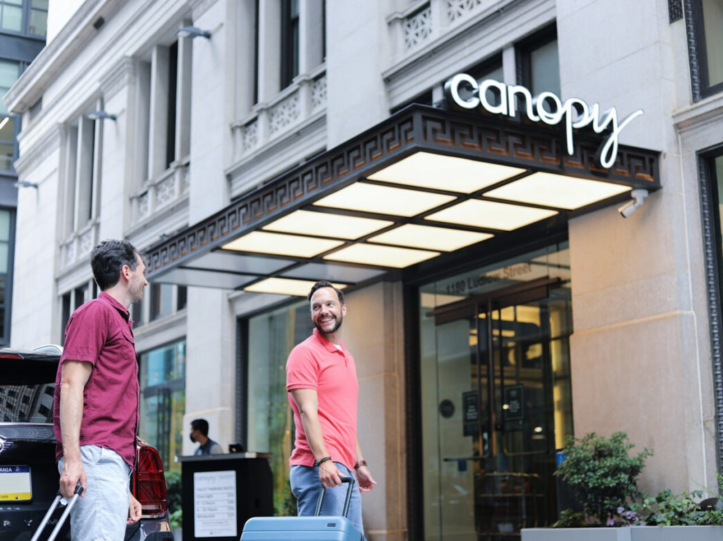 Two people wheel their luggage toward the entrance of the Canopy by Hilton hotel.