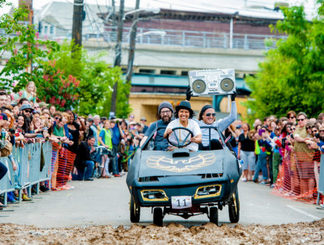 Three people pedal a people-powered car toward a mud pit at the Kensington Derby & Arts Festival
