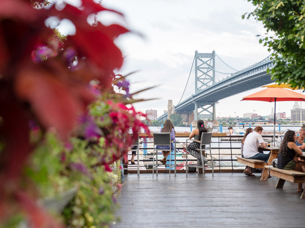 People sit at long tables and bar stools looking at the Benjamin Franklin Bridge at Morgan's Pier in Philadelphia