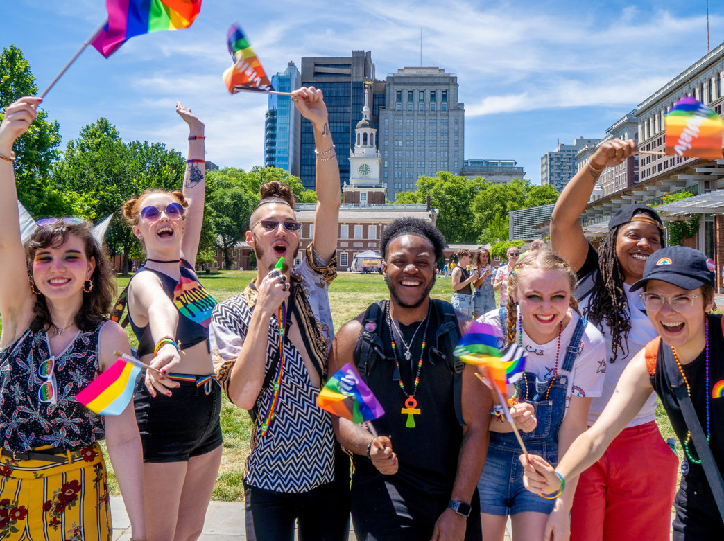Six people wearing Pride-related clothing pose on Indepdnence Mall with Independence Hall in the background in Philadelphia