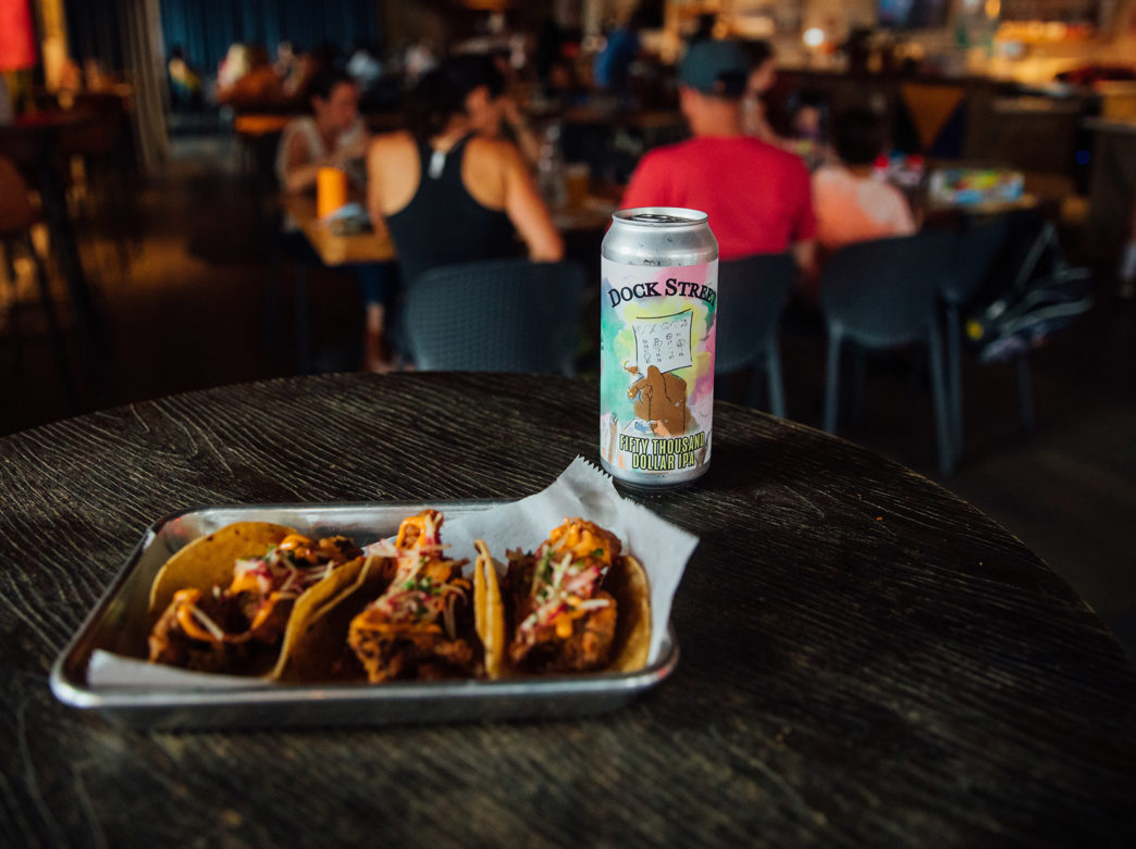 An image of food on a plate next to a beer with people in the background at Dock Street Brewery
