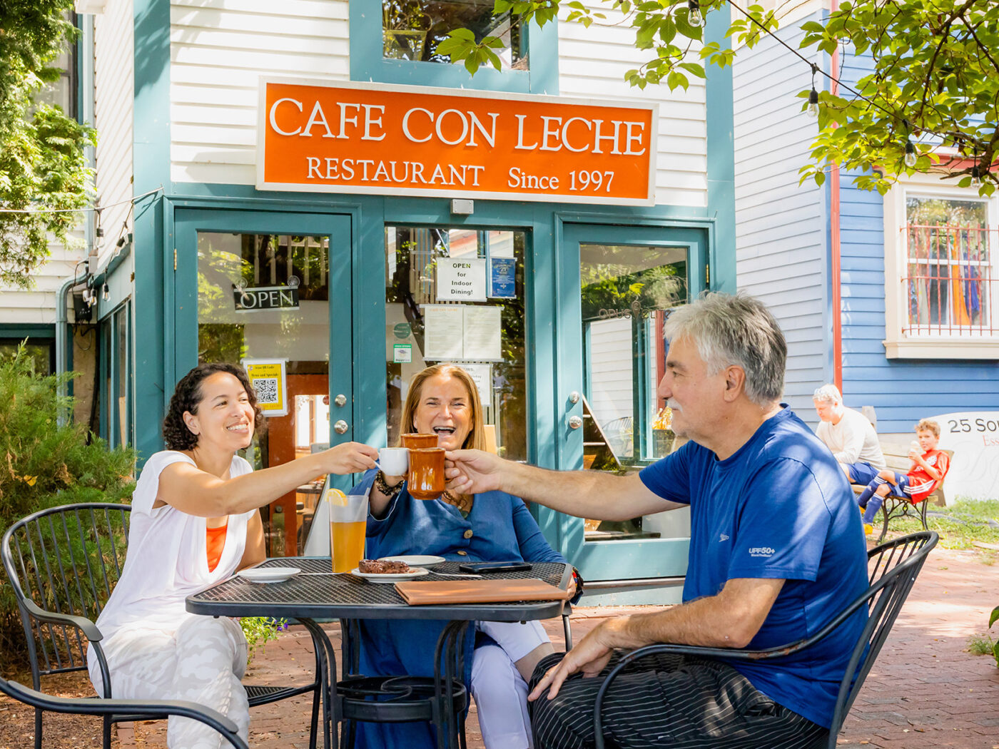 Three diners seated at an outdoor table toast with coffee mugs at Cafe Con Leche in Newtown
