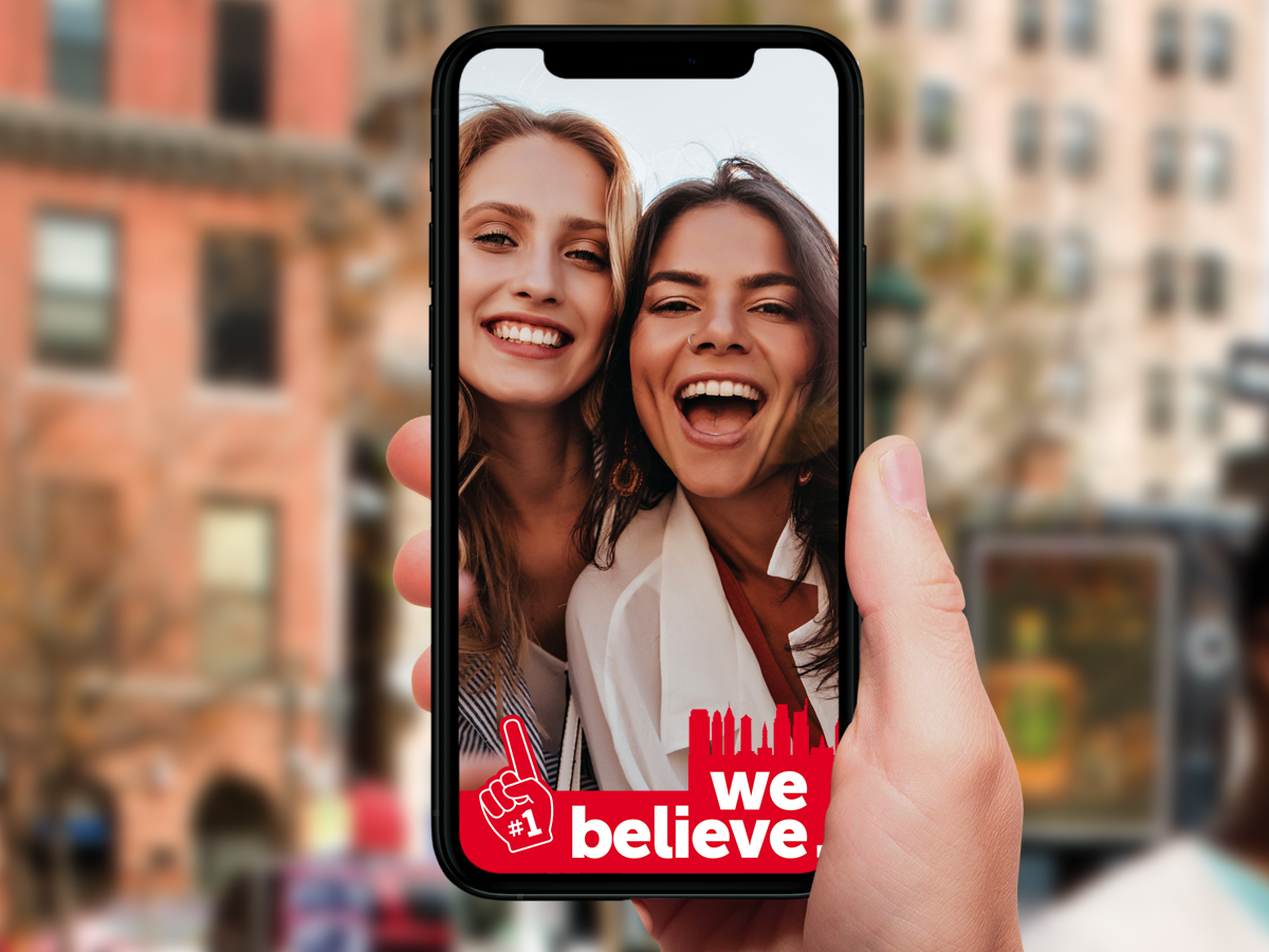 A hand holds a phone with Visit Philadelphia's We Believe filter in front of two people smiling