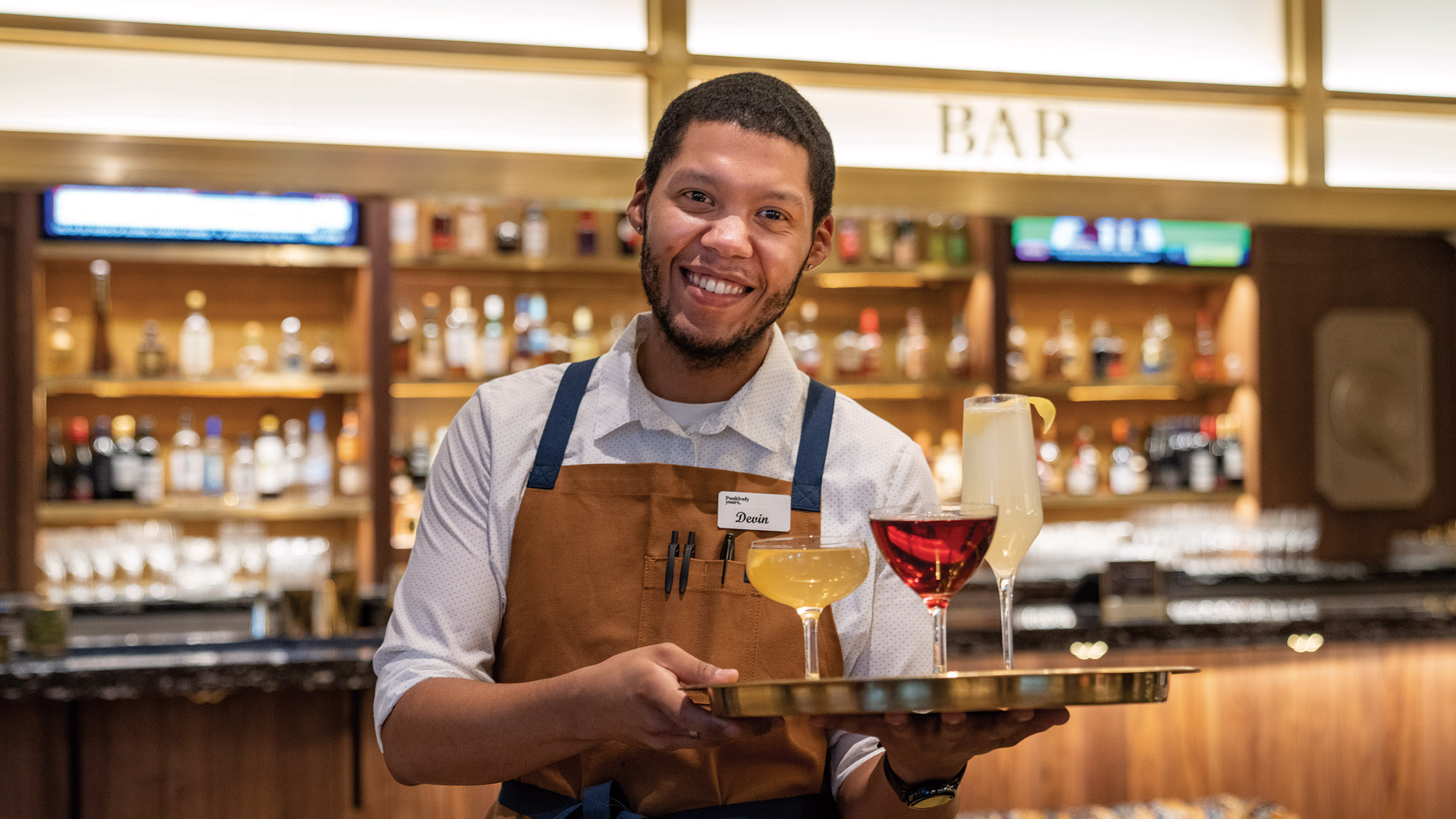 A waiter holds a tray of drinks and smiles at the camera