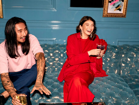 Two people sit on a blue/green sofa holding drinks at laughing at Stevens' Prophecy inside the W Philadelphia