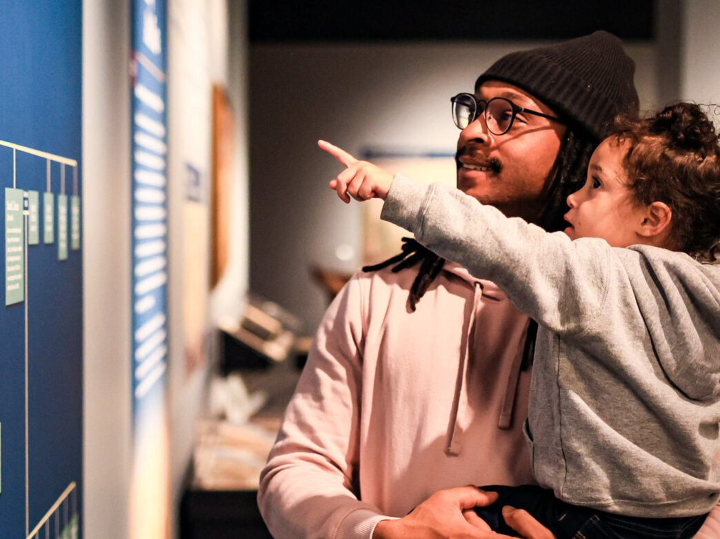 A father holds his daughter as she points at a museum exhibit at the Museum of the American Revolution