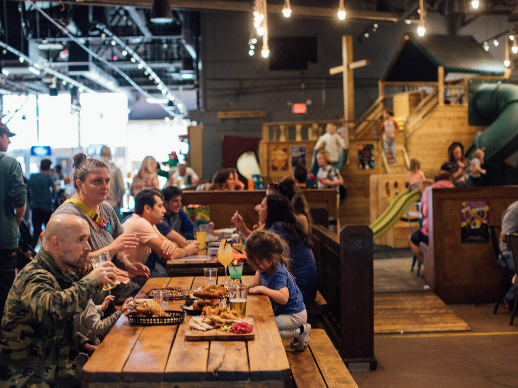 People eat at a wooden table with an indoor playground in the background at Craft Hall in Philadelphia