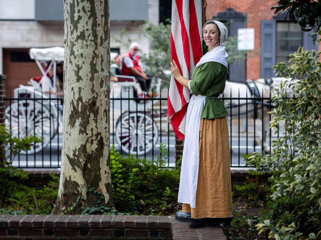 A re-enactor dressed as Betsy Ross poses next to an American flag.