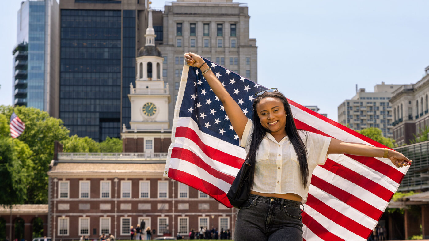 A person stands facing the camera holding an American flag behind their back. In the background is Philadelphia's Independence Hall.