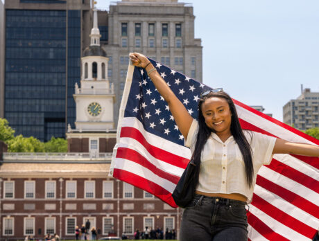 A person stands facing the camera holding an American flag behind their back. In the background is Philadelphia's Independence Hall