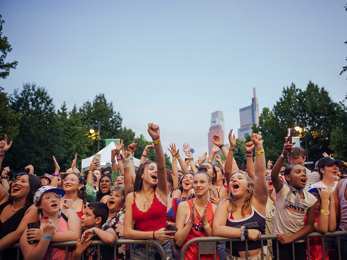 A crowd of people with their hands in the air awaiting the Fourth of July concert on the Ben Franklin Parkway.