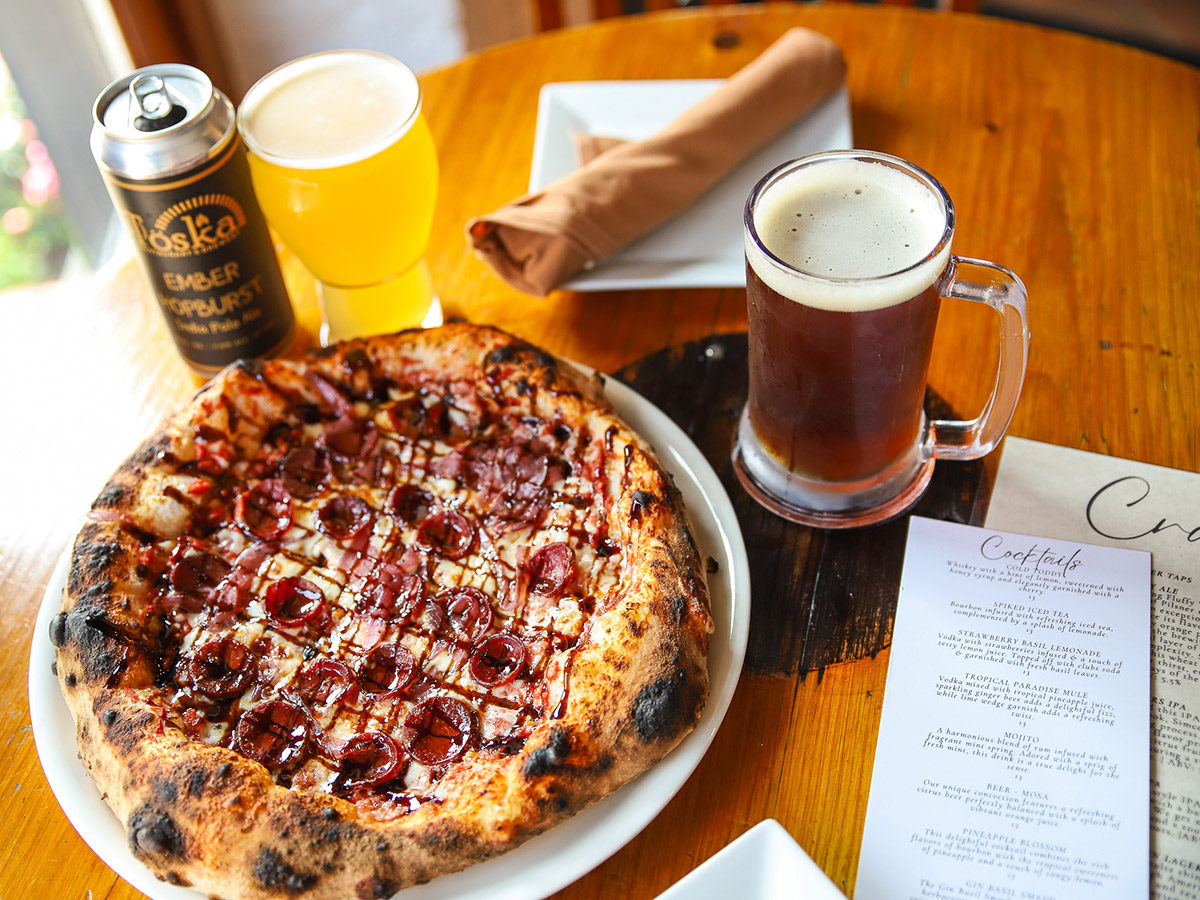 A pepperoni pizza sits on a table next to two glasses of beer. A can on the table is labeled "Toska Ember Hopburst."