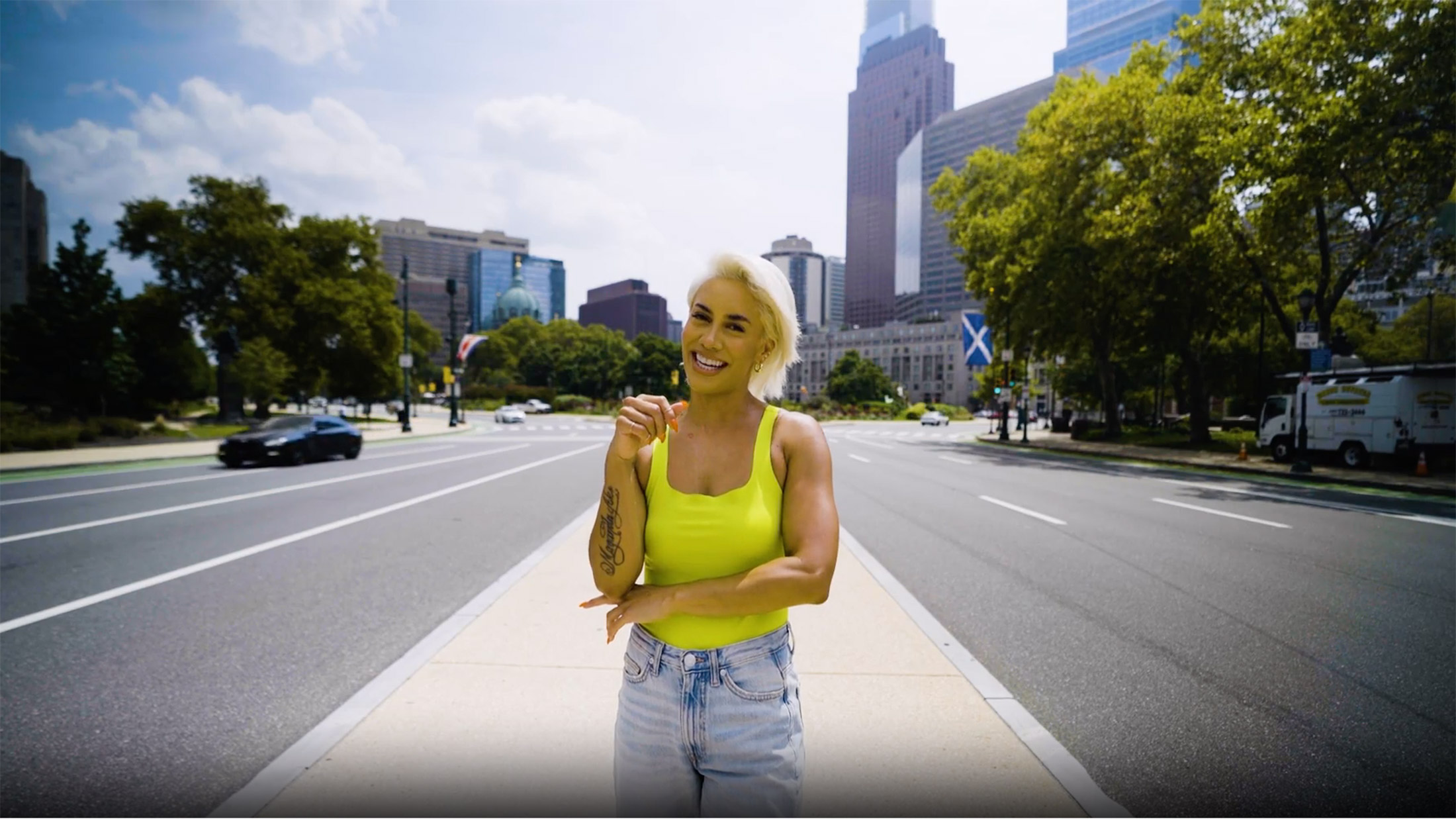 A person wearing a yellow top and jeans smiles while standing on a median on the Benjamin Franklin Parkway in Philadelphia