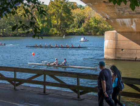 People walk along the Schuylkill River Trail while rowers during the Head of the Schuylkill Regatta row past in Philadelphia
