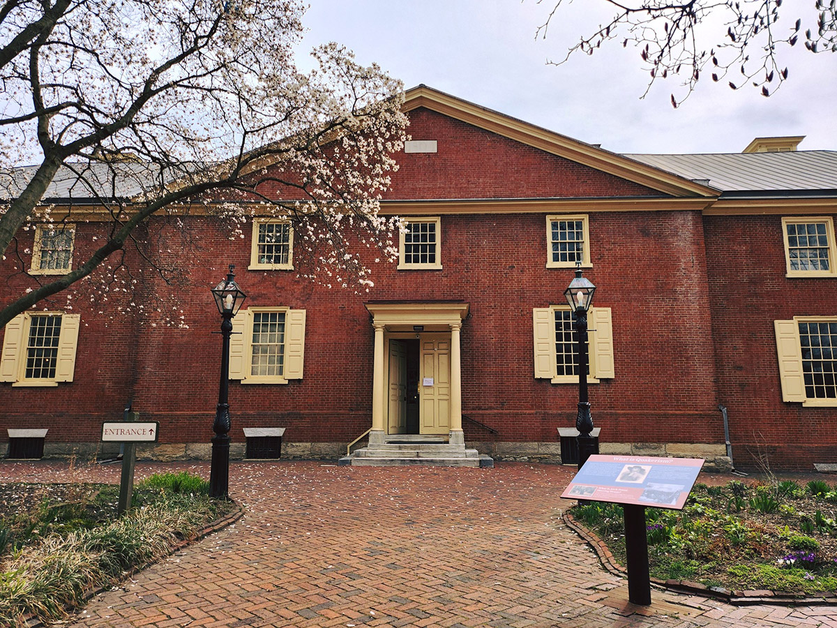 Entrée extérieure de l'Arch Street Meeting House, un bâtiment en briques rouges avec des encadrements de fenêtres et des volets de couleur crème, des poteaux d'éclairage et des panneaux de signalisation.