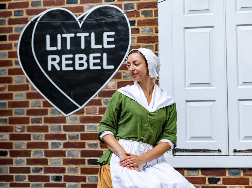 Young Betsy Ross sits in front of Betsy Ross House with a black heart painted on the building exterior with Little Rebel written inside the heart in white lettering.