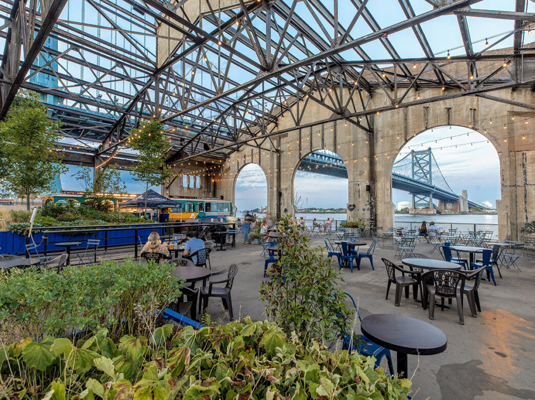 Outdoor dining tables, patio string lights and guests at the Garden at Cherry Street Pier overlooking the Delaware River during the summer.