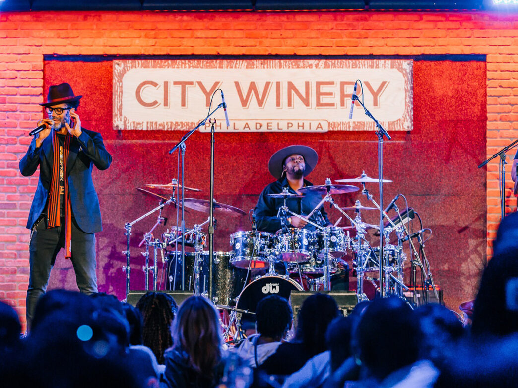 A band performs on stage with a City Winery banner hanging behind the band. The lead singer sings into the microphone and the drummer plays behind the singer.