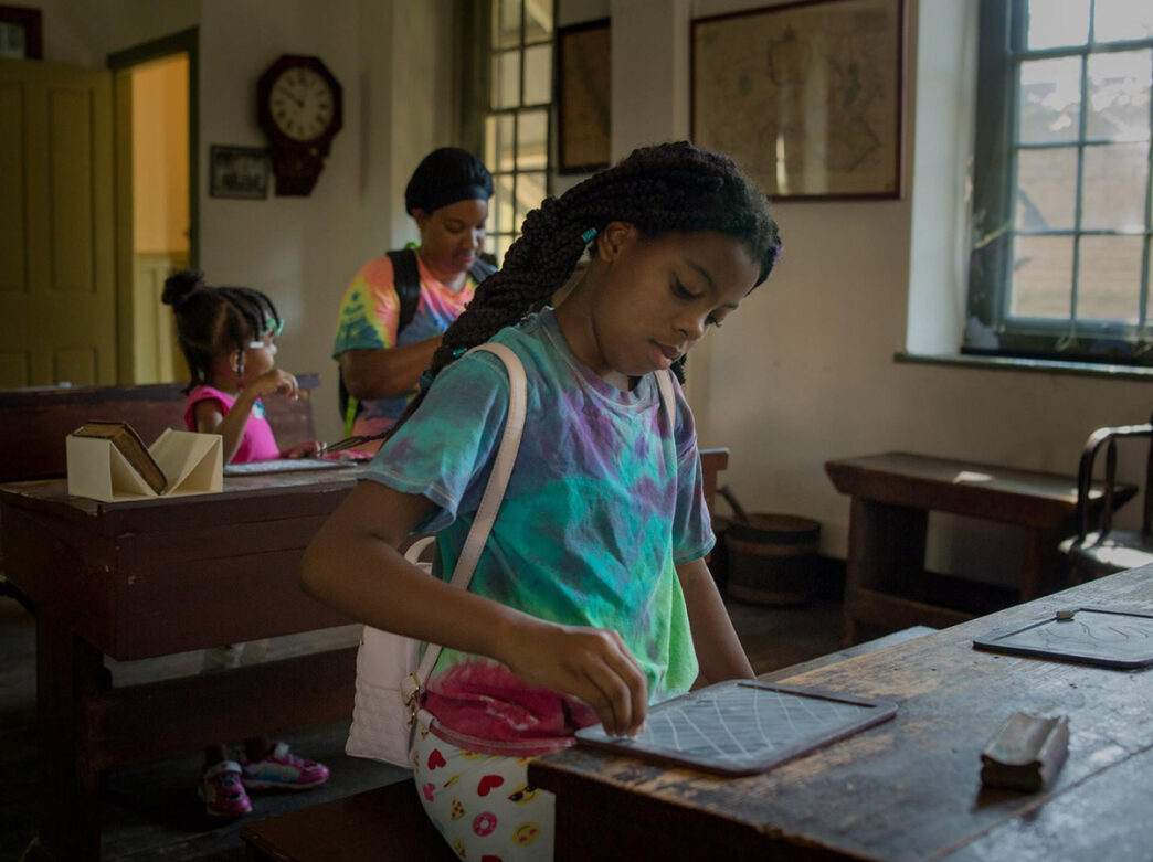 Child drawing on small chalkboard at a desk inside the Concord School House.