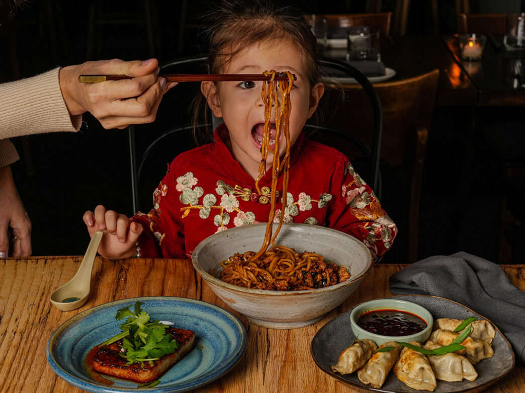 Child opens mouth wide to eat Chinese noodles hanging in front of them on chopsticks held by parent.