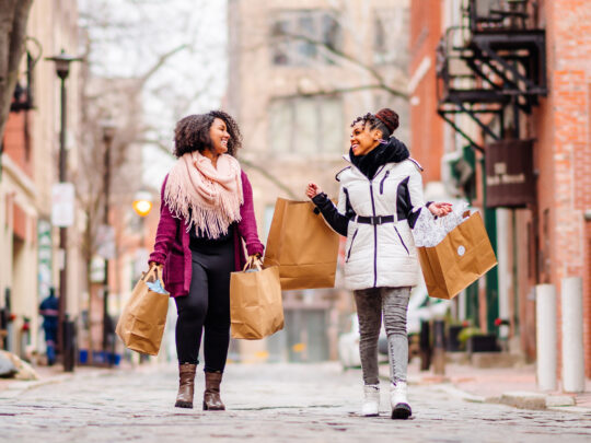 Duas mulheres caminham por uma rua de paralelepípedos na Filadélfia Old City com várias sacolas de compras nas mãos.