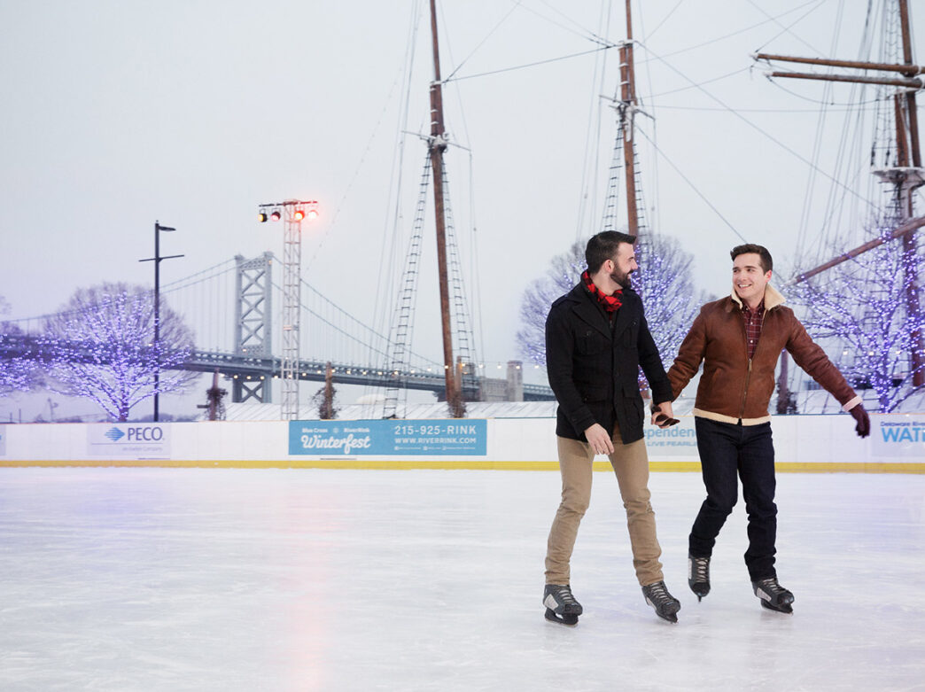 Two men wearing winter jackets smiling, holding hands, and skating across the ice skating rink at Independence Blue Cross RiverRink Winterfest. Trees with twinkling lights and the Benjamin Franklin bridge can be seen in the background behind the couple.
