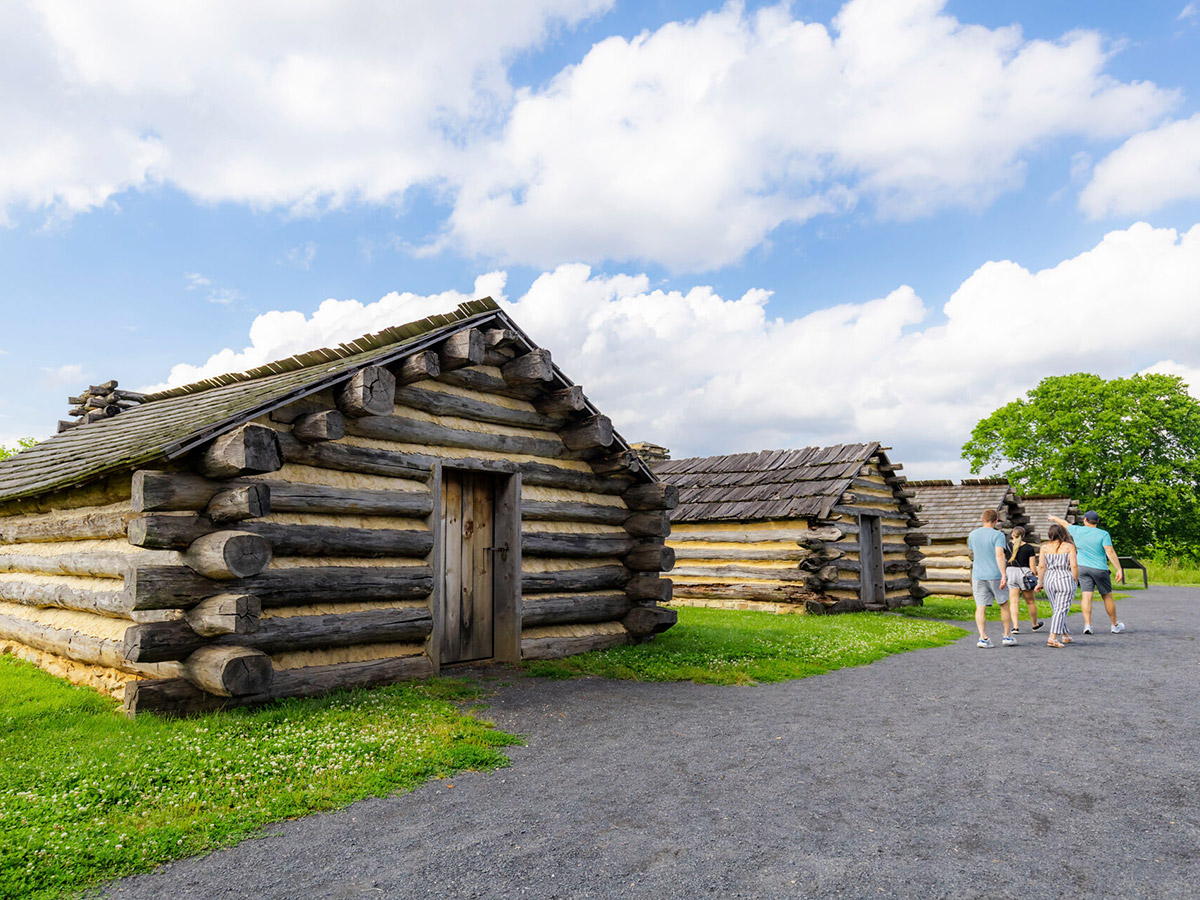 Group of four adults walking on a gravel path with reconstructed log soldiers' huts at Valley Forge National Historical Park on a summer day.