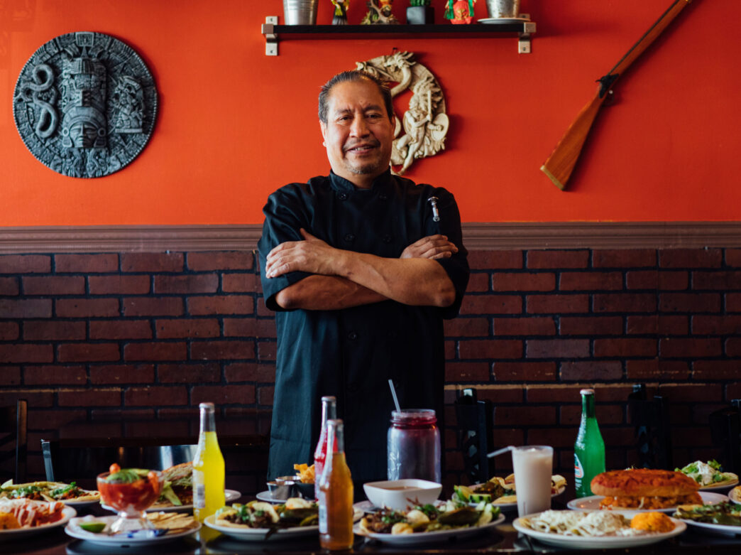 A chef wearing all black stands with his arms crossed behind a table with drinks and meals at Adelitas.