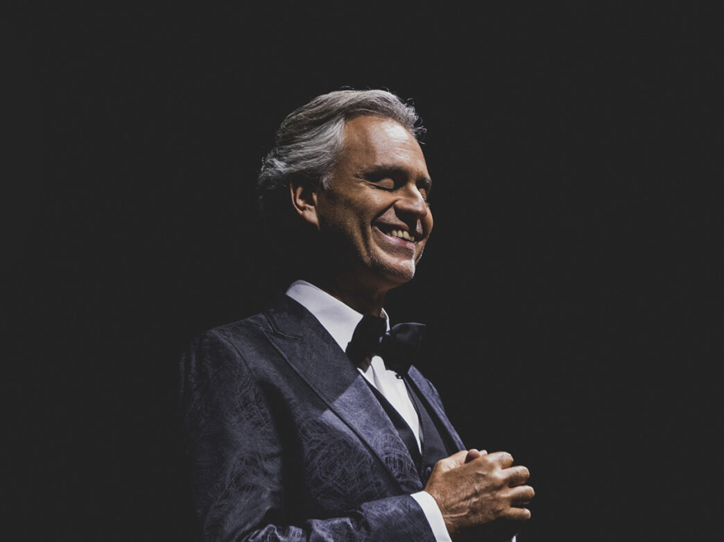 Andrea Bocelli stands with his hands at his chest in front of a black background while wearing a suit and bowtie.