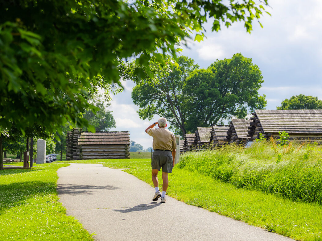 Man in shorts walks away from camera along a paved path lined with green trees and historic cabins at Valley Forge National Historical Park.