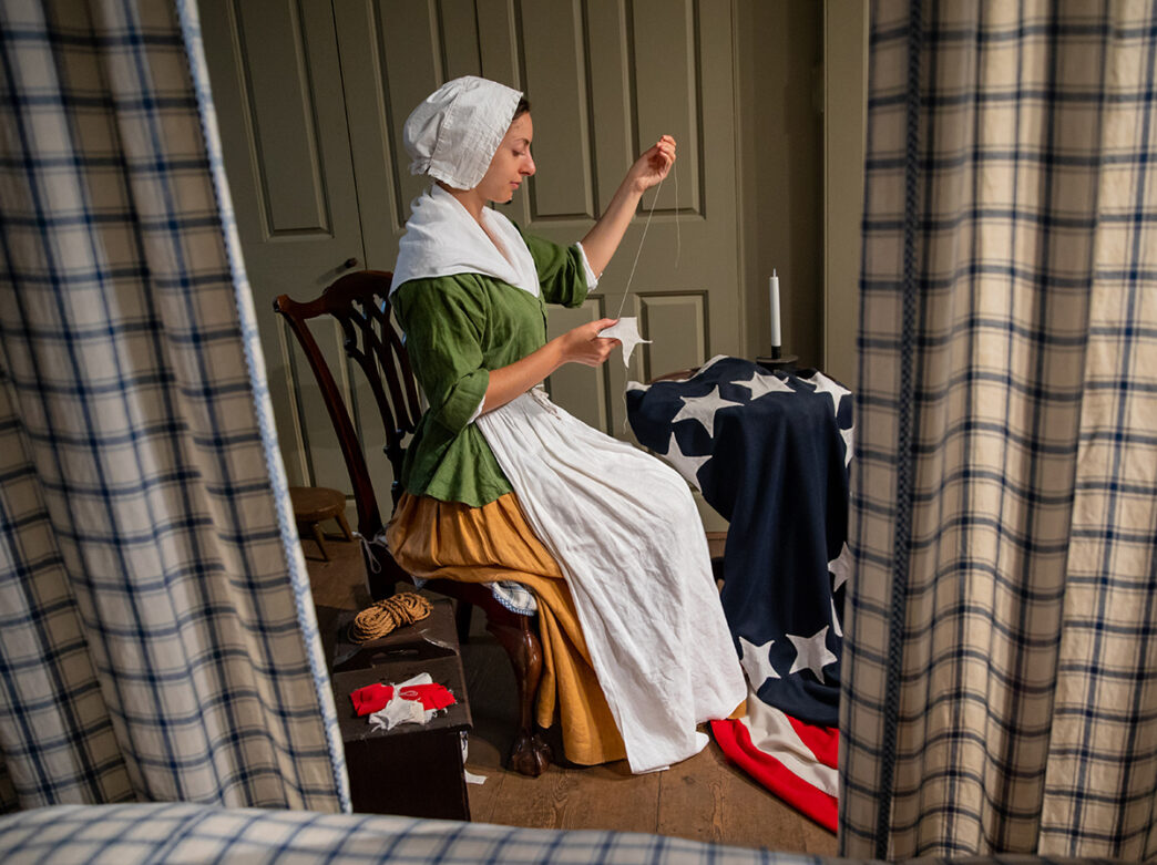 Um reencenador interpretando Betsy Ross está sentado em uma cadeira costurando uma estrela branca para ser costurada em uma bandeira americana.