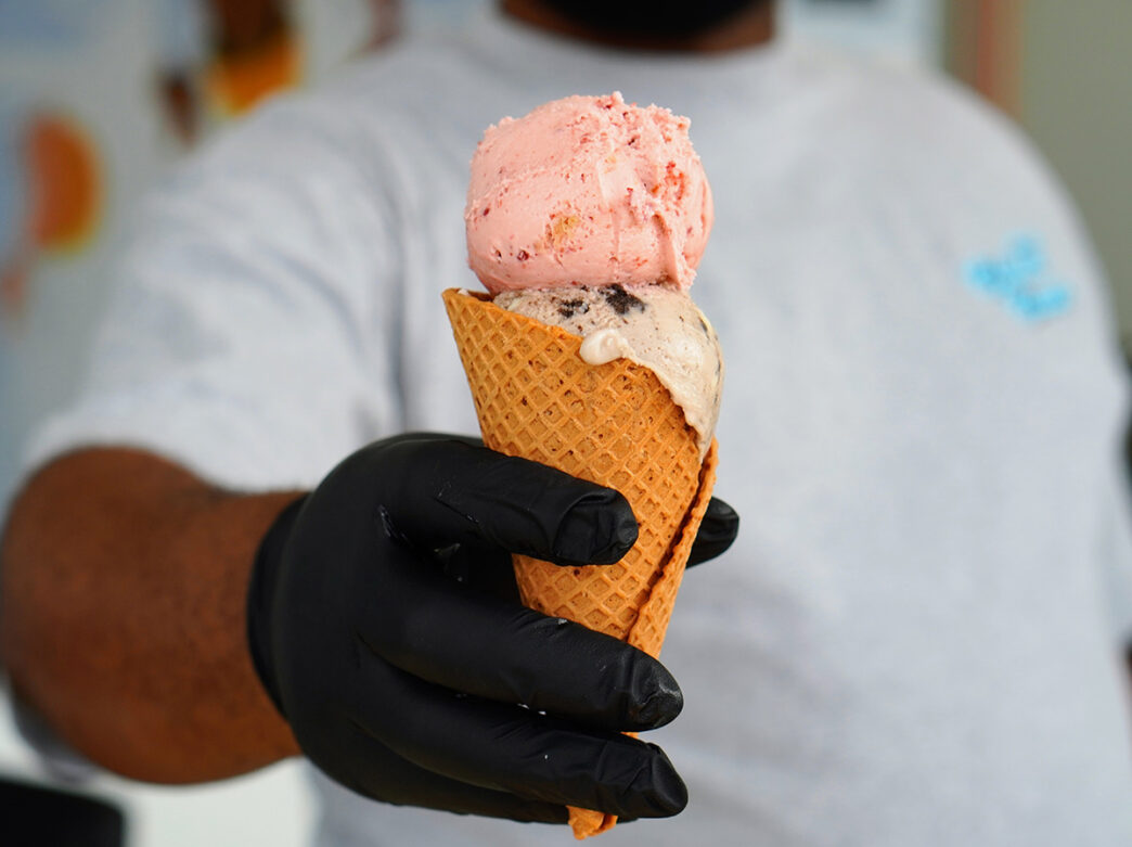 A server wearing a black glove holds out a cone with two scoops of gelato to the camera at Cloud Cups in Philadelphia.