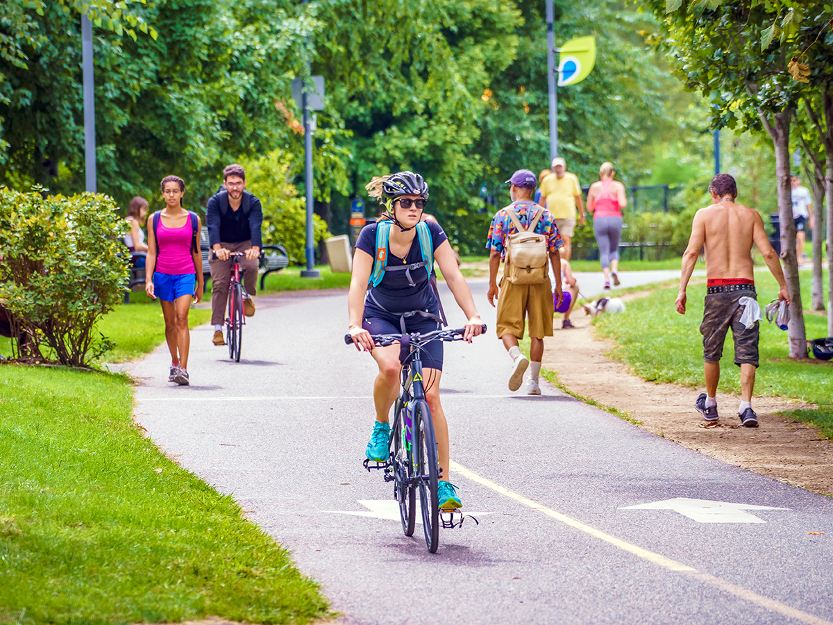 A biker wearing a helmet and teal sneakers rides along the Schuylkill River Trail amongst other bikers and walkers.