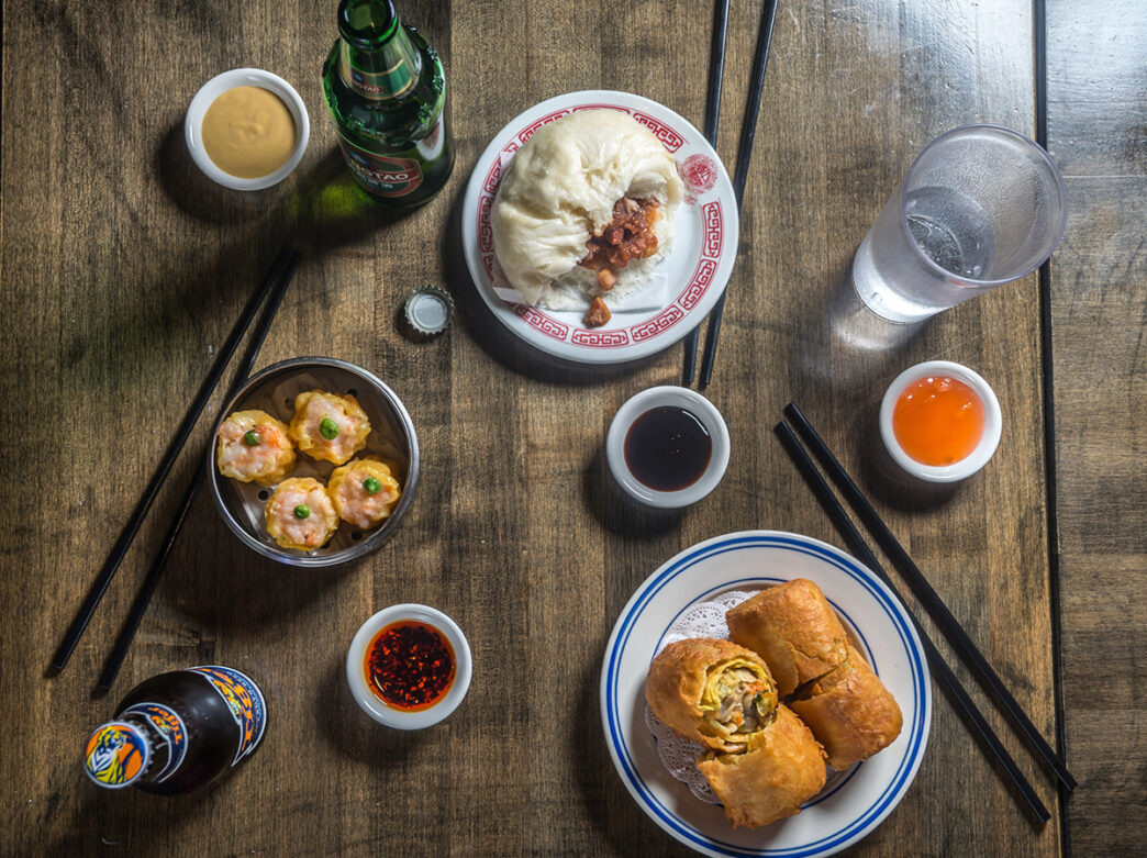 Assorted menu items including egg rolls, shrimp sui mai and a roast pork bun from Nom Wah in Philadelphia are displayed on a wood table.