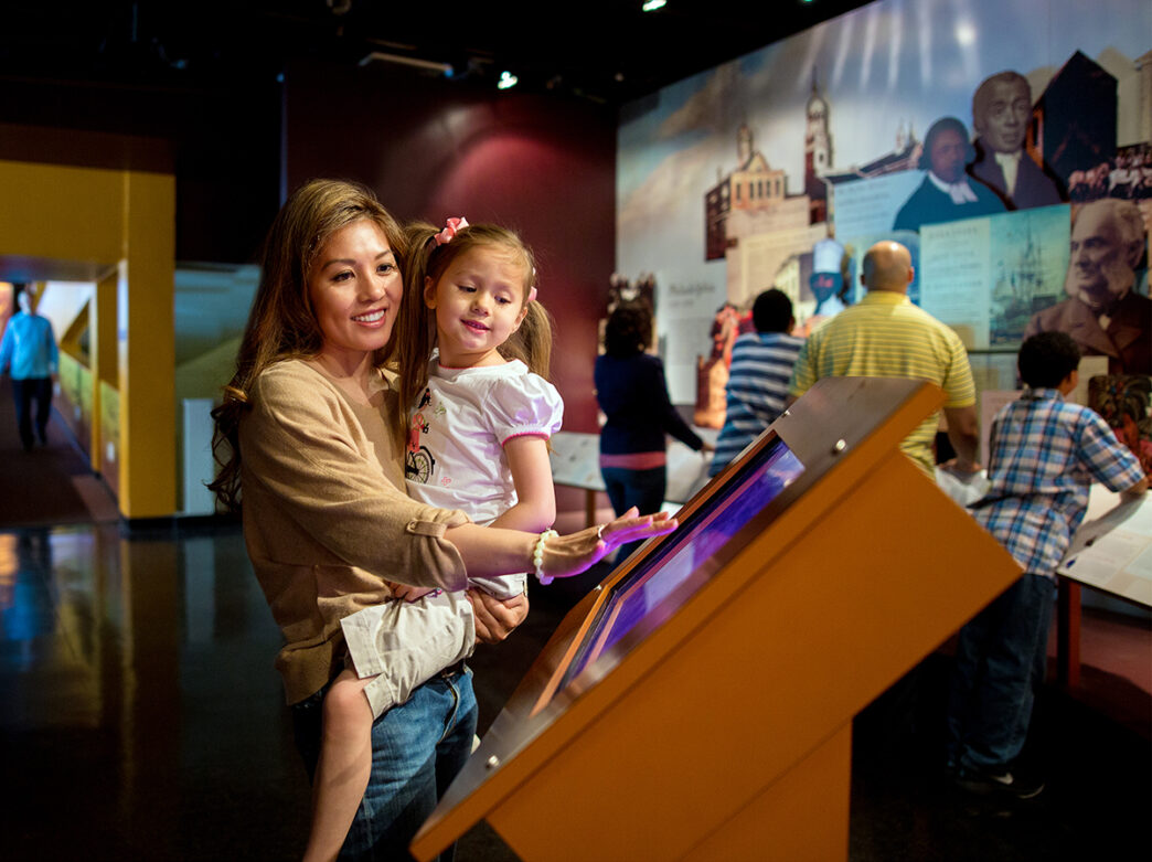 A woman holds a child while she touches an interactive screen at an exhibit at the African American Museum in Philadelphia.