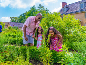 Een vader en twee dochters verkennen de tuinen van Bartram's Garden in Philadelphia. Eén kind buigt voorover om aan een bloem te ruiken.