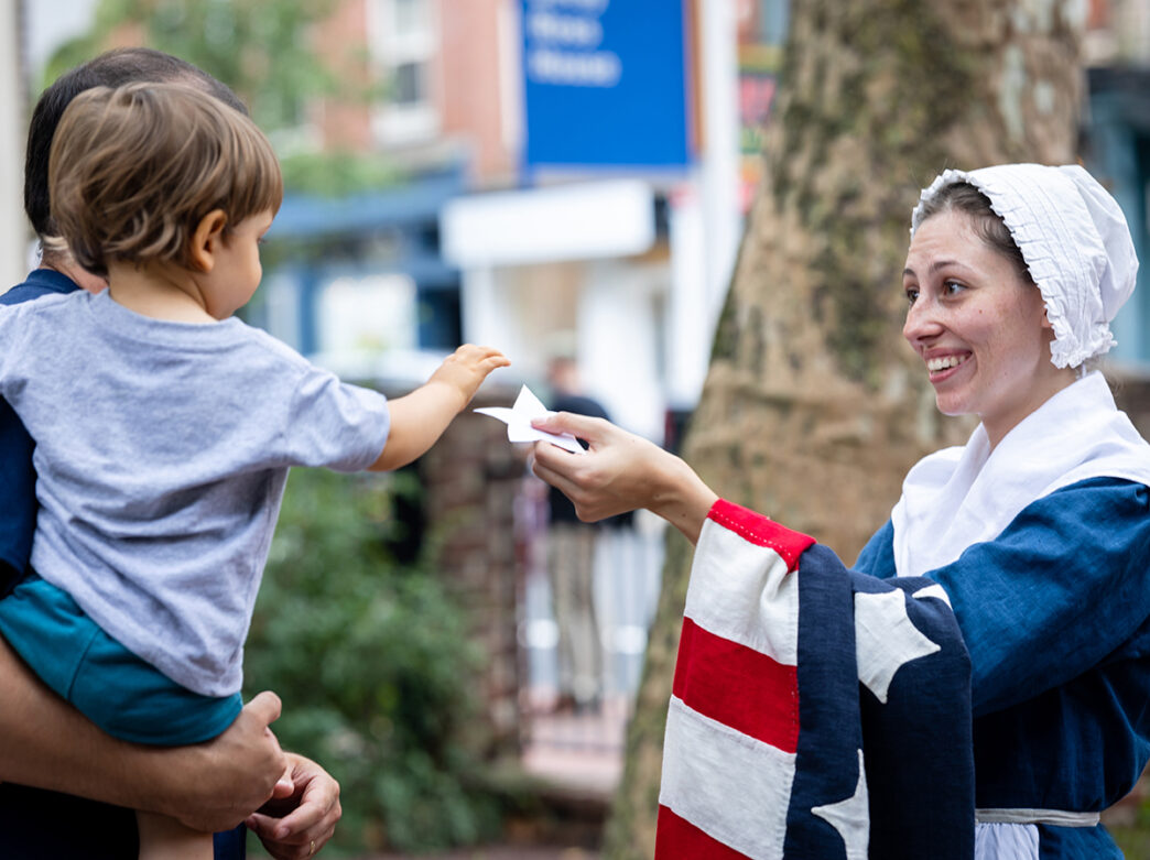 A reenactor playing Betsy Ross hands a white star to a child being held by their parent.