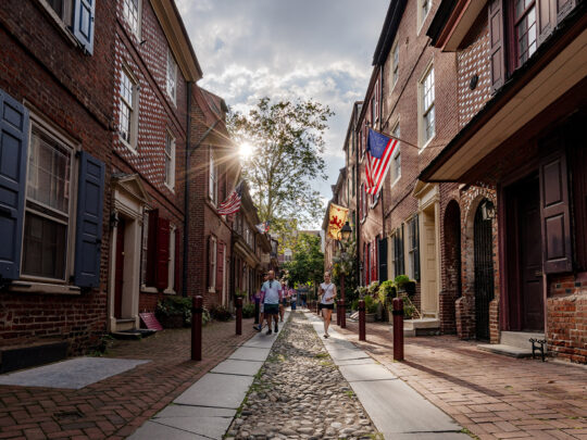 People walk along the cobblestone streets of Elfreth's Alley in Philadelphia with the sun rising up from behind a house.