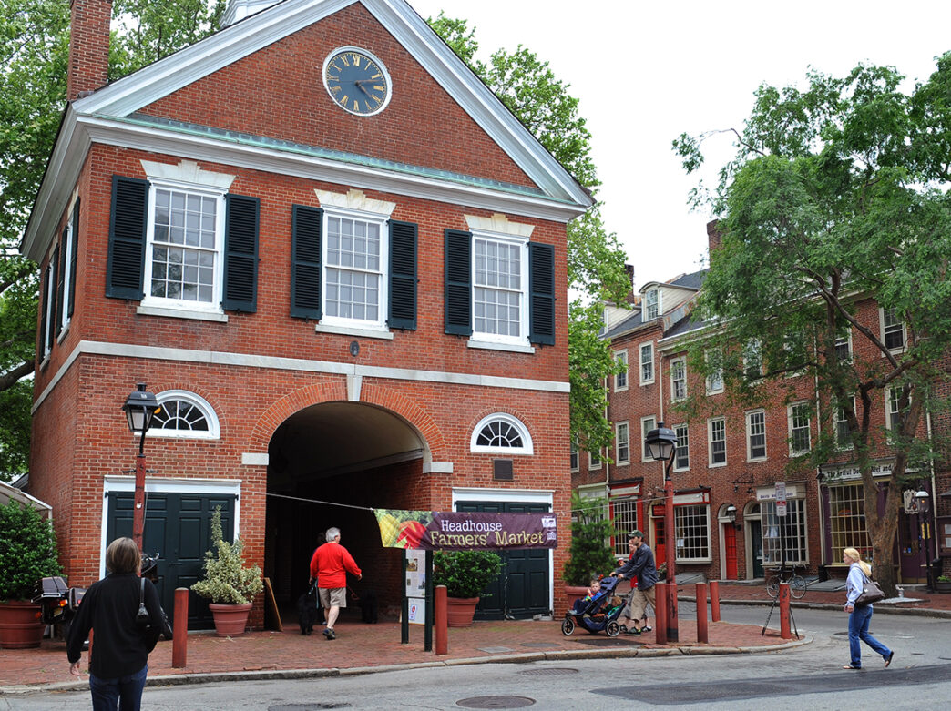 People walk through the street to enter a brick building with 3 windows and a clock on the exterior. The building hosts the Headhouse Farmers' Market.